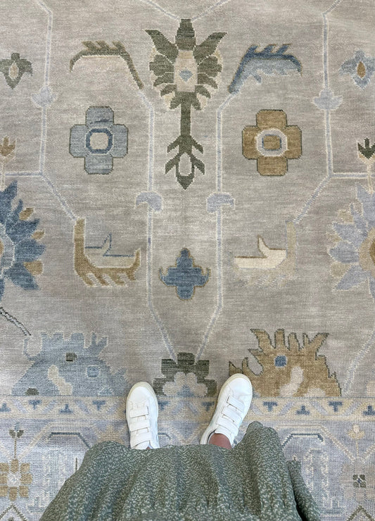 A top-down view of a person standing on a large patterned area rug, with their white sneakers and dark green textured skirt visible at the bottom edge.