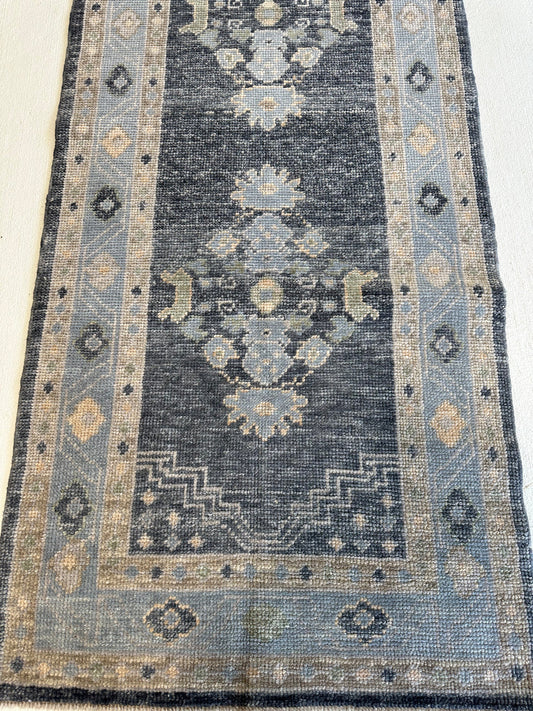 A rectangular area rug with a distressed traditional design, featuring a dark charcoal gray center field, light blue-gray borders, and beige accents.