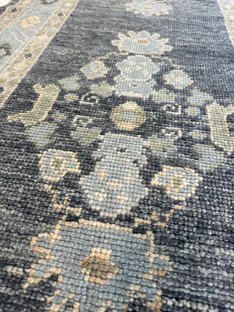 Close-up of a traditional patterned rug with a dark charcoal gray base and beige, cream, and muted blue-gray floral motifs.