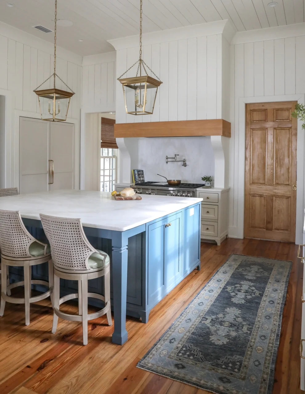 A bright kitchen interior with a blue island, white shiplap walls, hardwood floors, two hanging lantern lights, two woven bar stools, a wood range hood, and a long blue-gray patterned rug.