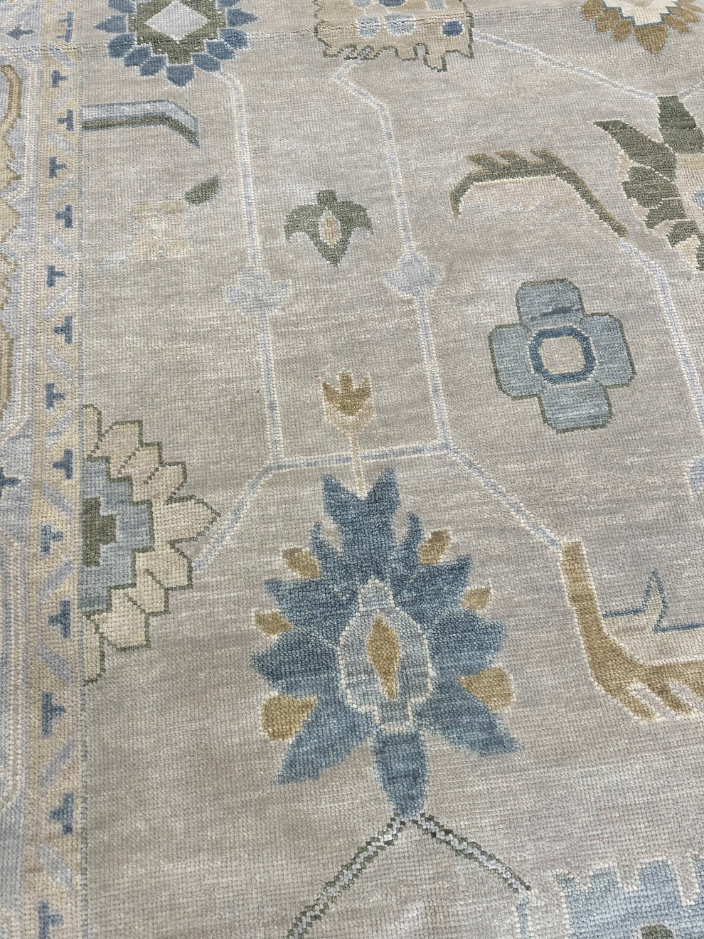 A close-up of a woven area rug with a traditional floral and geometric pattern in muted blue, beige, gray, and olive green tones.