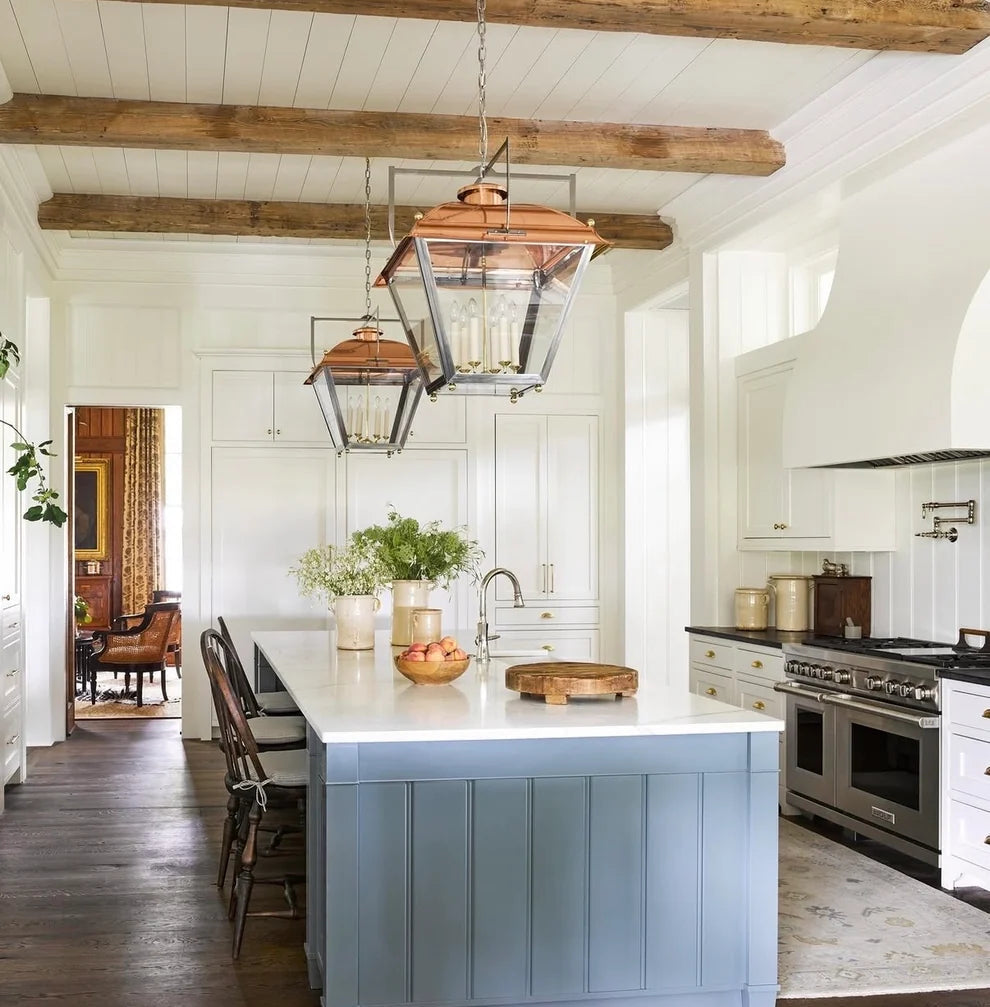 A bright, modern kitchen interior with a light blue paneled island, white shiplap walls, exposed wooden ceiling beams, and two copper-and-glass pendant lights hanging above the island. A visible 'verandamag' logo appears at the top of the image.