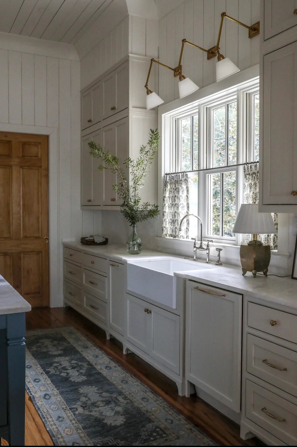 A bright, modern farmhouse-style kitchen featuring white shaker-style cabinets, a white farmhouse sink, brass wall sconces, and a blue patterned rug on a hardwood floor.
