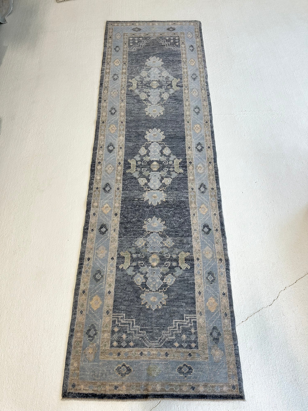 A long rectangular area rug laid flat on a light concrete floor, featuring a distressed traditional design with a dark gray center, beige and light blue-gray borders, and faded floral medallions.