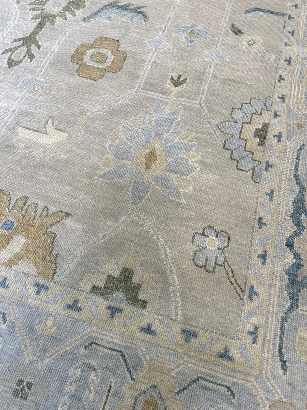 A close-up of a traditional patterned rug with a light beige base and muted blue, gray, and tan floral motifs.