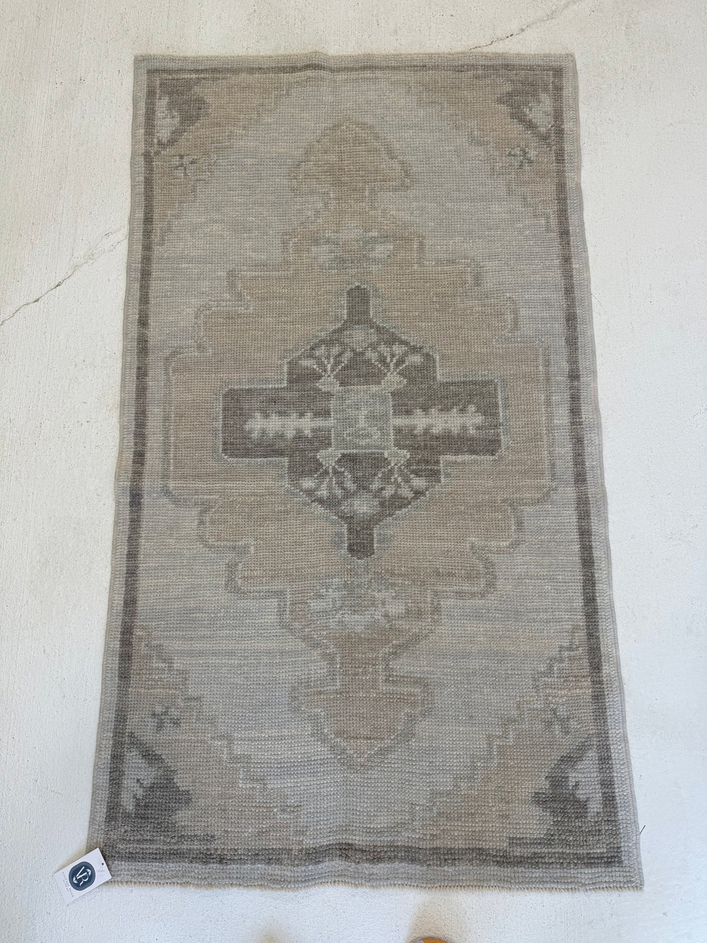 A rectangular vintage-style area rug with a distressed geometric medallion pattern in muted beige, gray, and brown tones, laid flat on a light off-white textured floor.