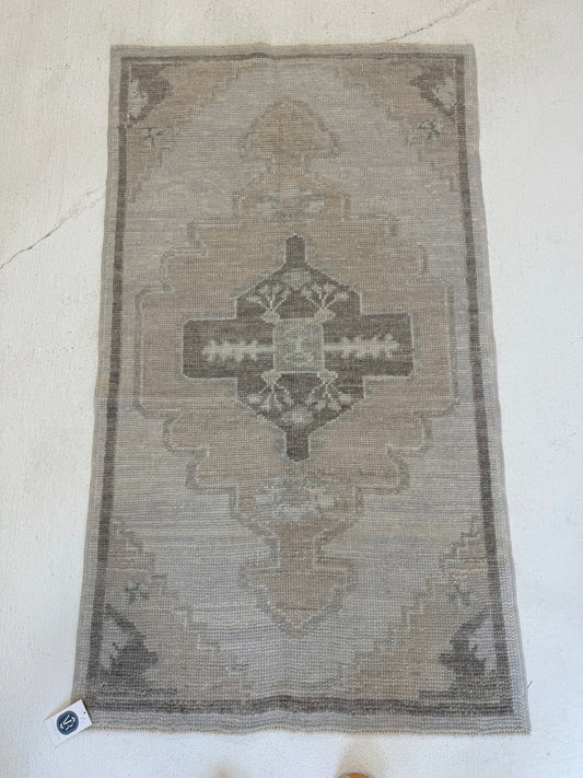 A rectangular vintage-style area rug with a distressed geometric medallion pattern in muted beige, gray, and brown tones, laid flat on a light off-white textured floor.