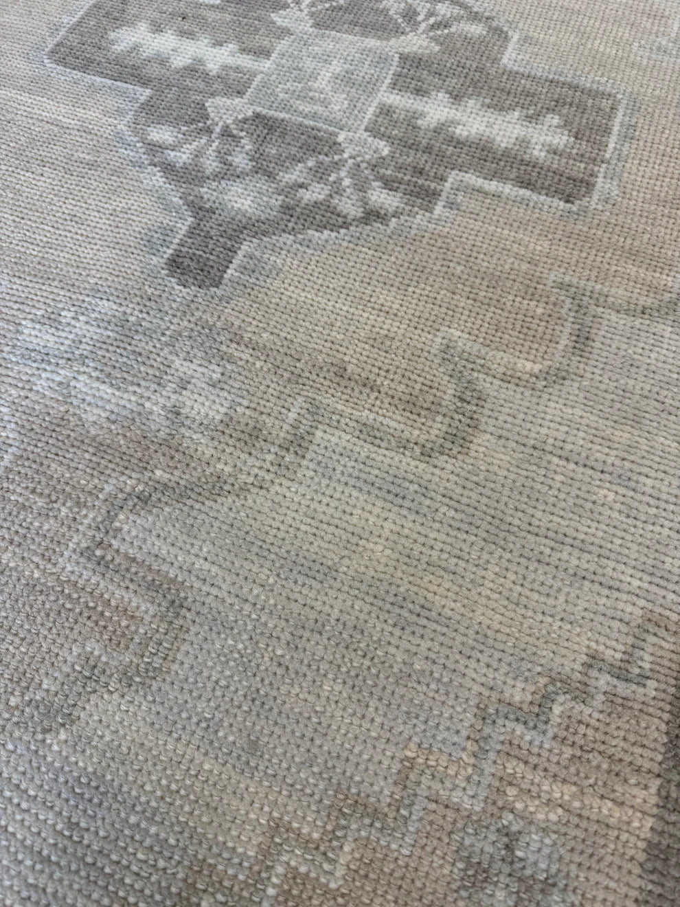 A close-up of a woven rug or textile showing a distressed, vintage-style geometric pattern in muted beige, gray, and taupe tones.