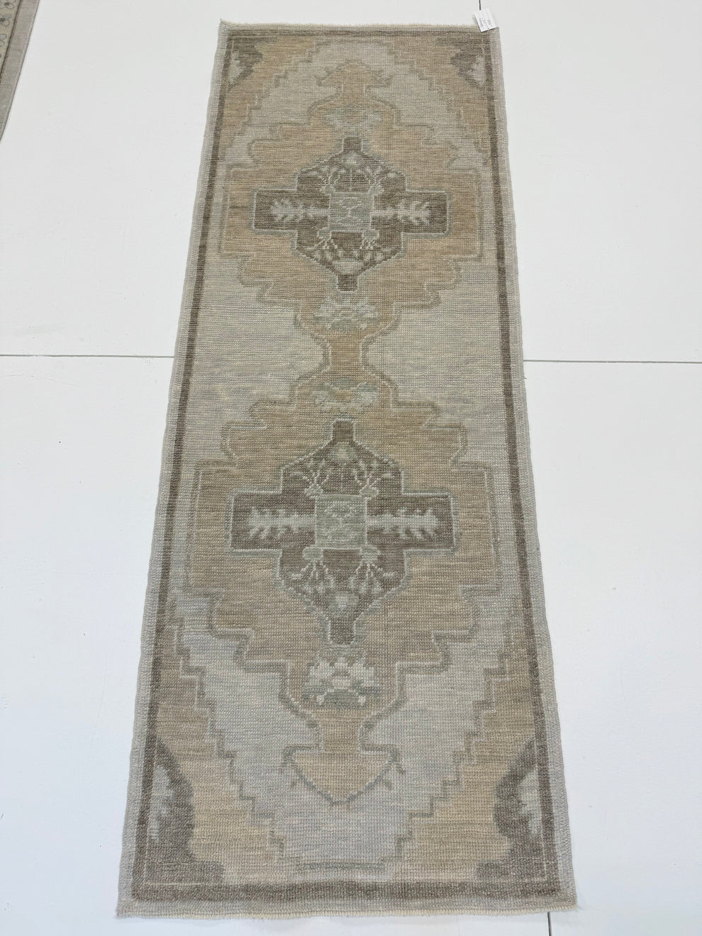 A long rectangular area rug laid flat on a white floor, featuring a distressed geometric and medallion pattern in muted beige, gray, taupe, and brown tones.