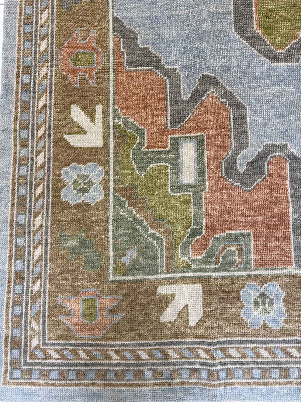 Close-up of a woven rug corner showing a geometric pattern with muted blue, brown, terracotta, olive green, cream, and light blue details.