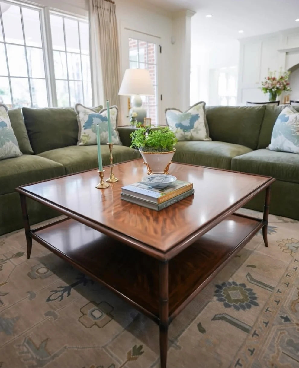 A dark wood coffee table with a lower shelf is styled in a bright living room with an olive green sectional sofa, patterned throw pillows, and a beige rug.