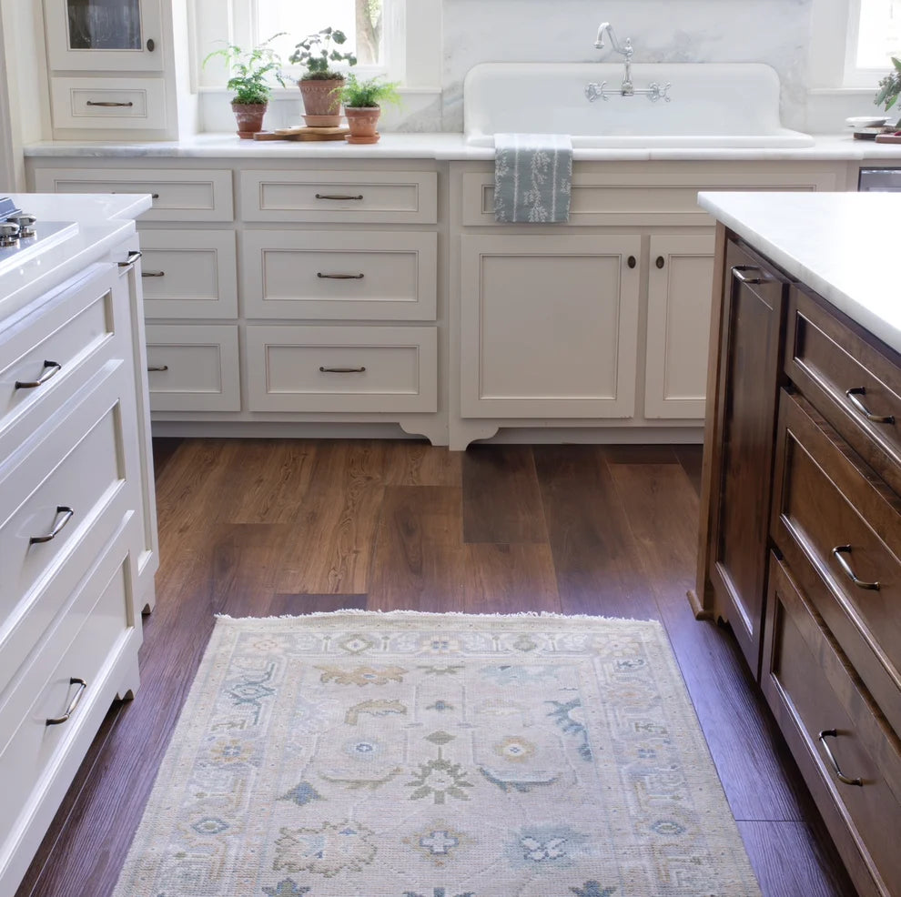 A rectangular area rug with a traditional pattern in beige, blue, and cream, placed on dark wood flooring in a bright kitchen with white and dark wood cabinets.