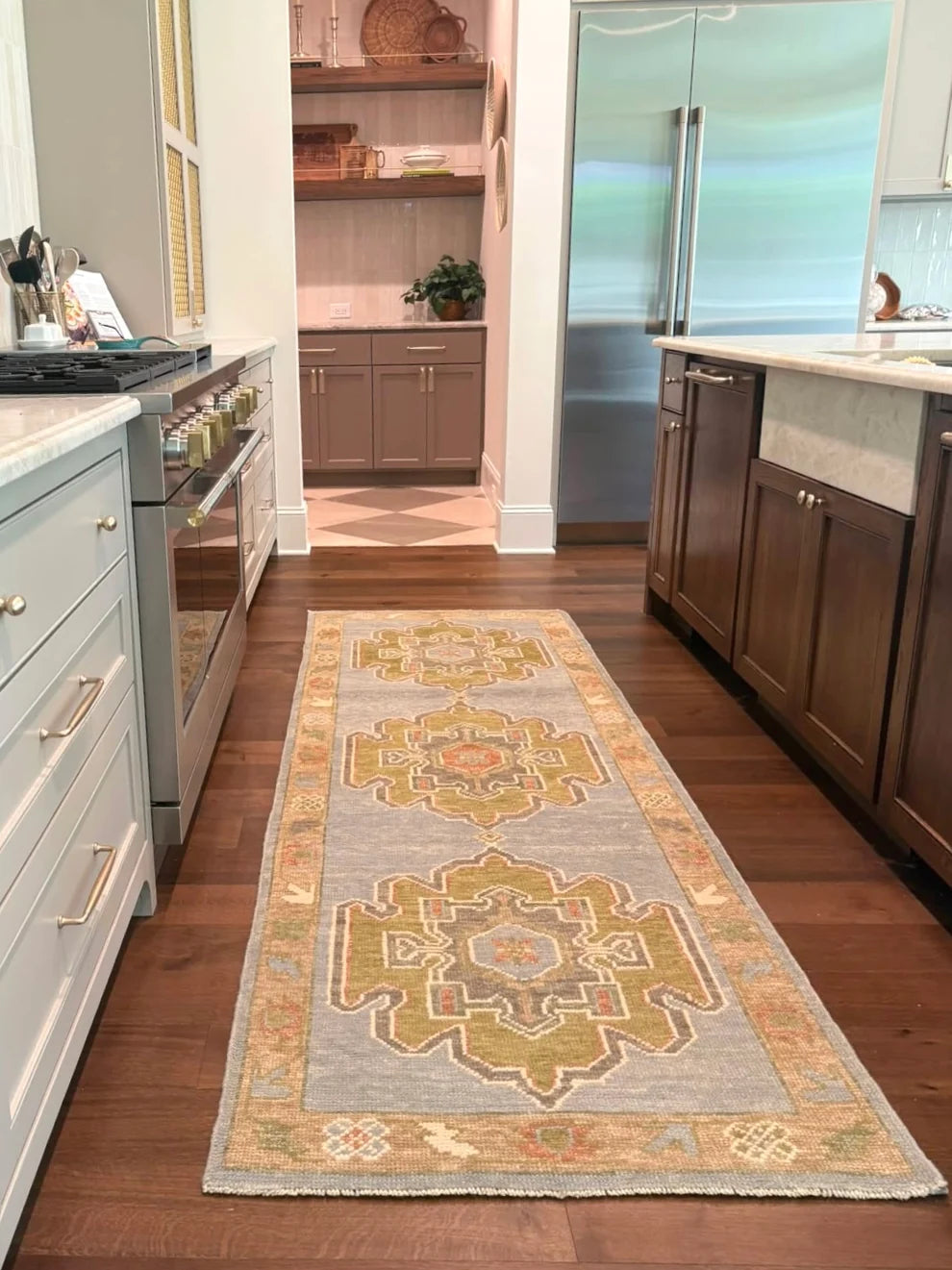A long rectangular area rug with a traditional medallion pattern in beige, gold, and muted orange on a light gray-blue background, shown on a dark wood kitchen floor between white and dark wood cabinets.