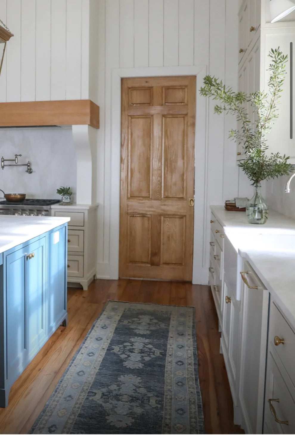 A rustic light brown wooden six-panel door set in a white paneled wall, flanked by a blue kitchen island on the left and white cabinets with gold handles on the right, with a patterned runner rug on the hardwood floor.