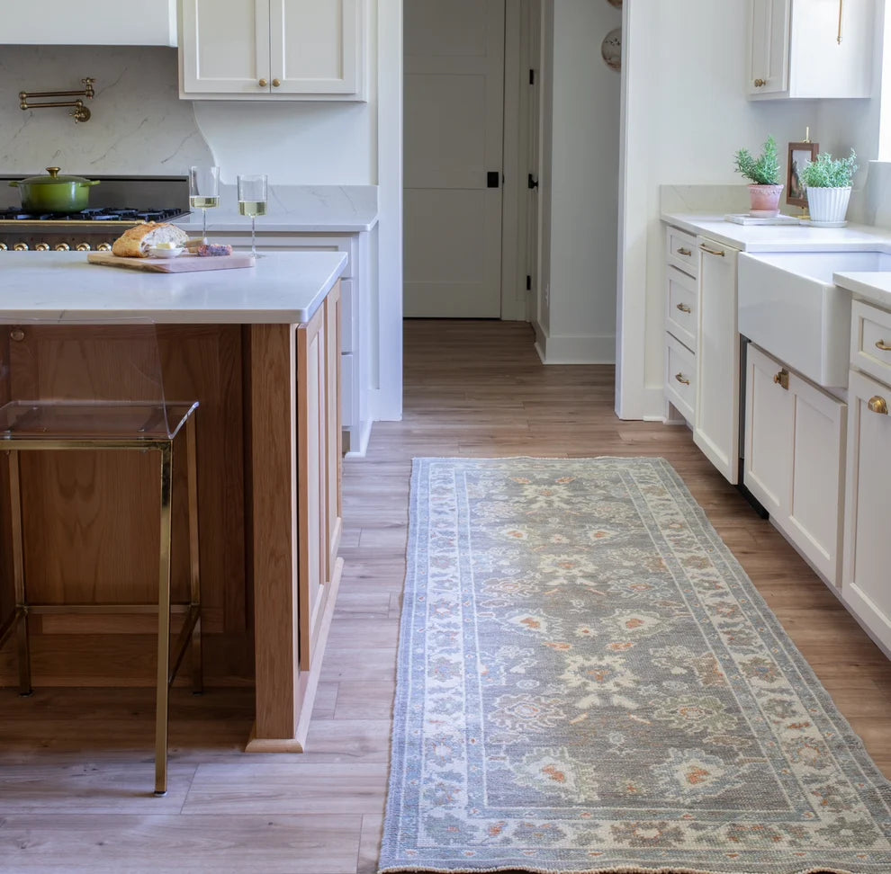 A long rectangular area rug with a traditional floral pattern in muted blue-gray, beige, and rust tones, shown on a light wood floor in a bright kitchen setting.