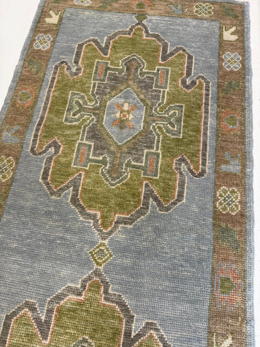 A flatweave area rug with a distressed geometric design, featuring a large central medallion in olive green and brown on a muted blue-gray field, with a patterned border.