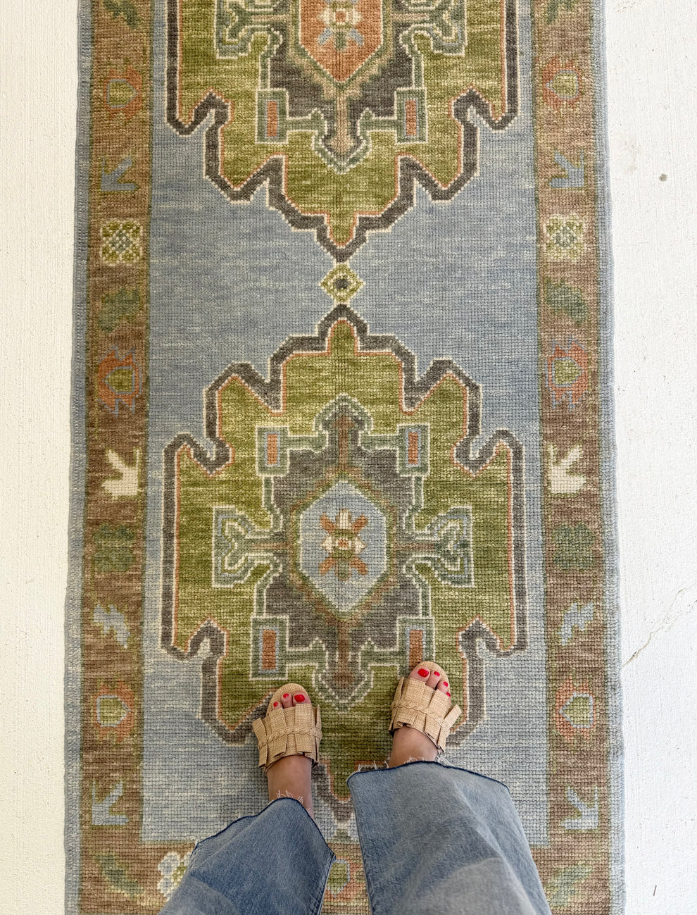 A top-down view of a long runner rug with a muted blue-gray field, olive green medallions, and brown decorative borders, shown on a white floor with a person's legs and sandals visible at the bottom.