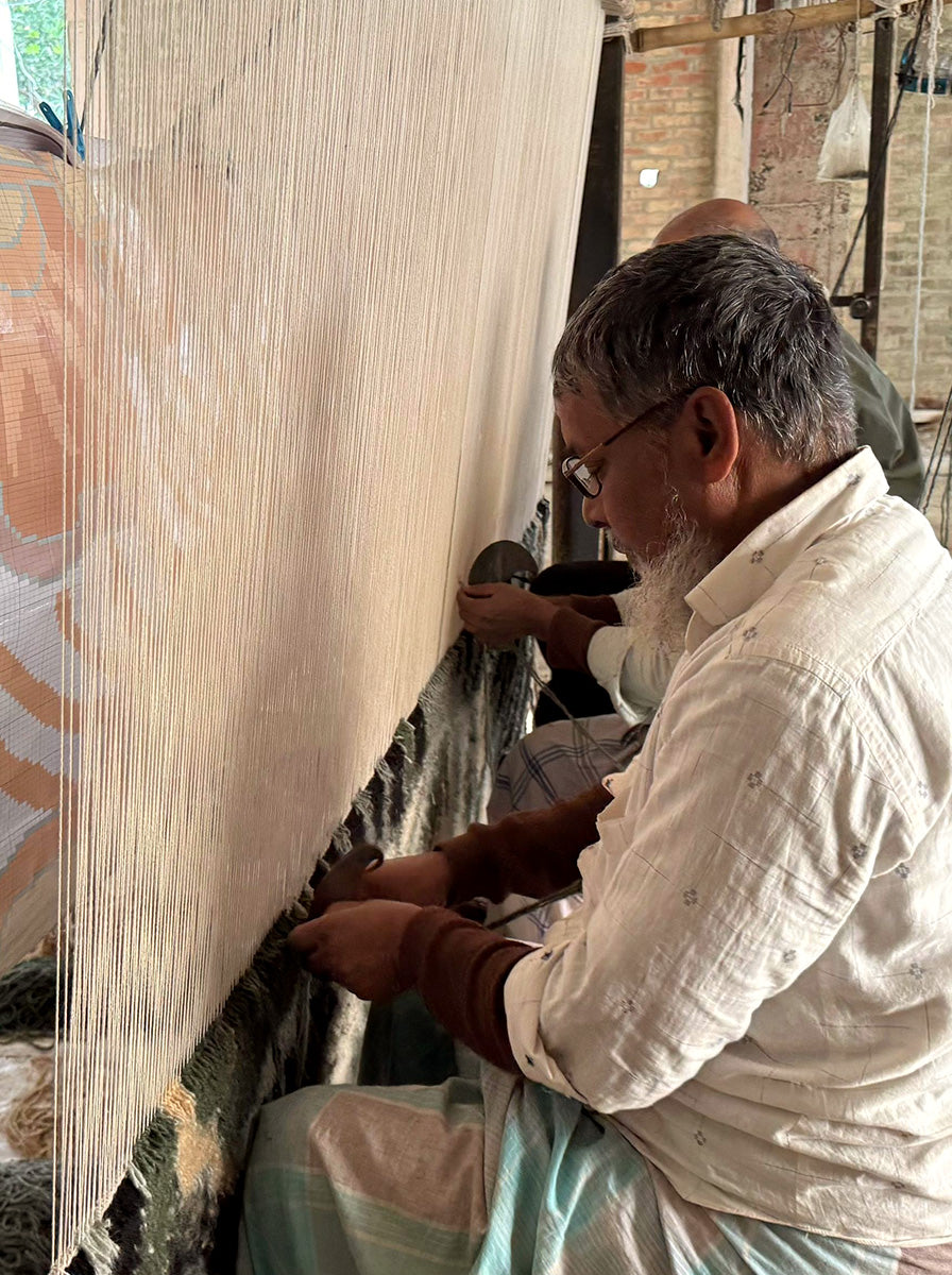 A weaver kneels beside a large loom, working on a patterned textile with off-white warp threads and a multicolored ground. The person wears a light cream shirt and a pale green-and-white drape, with workshop equipment and brick walls visible in the background.