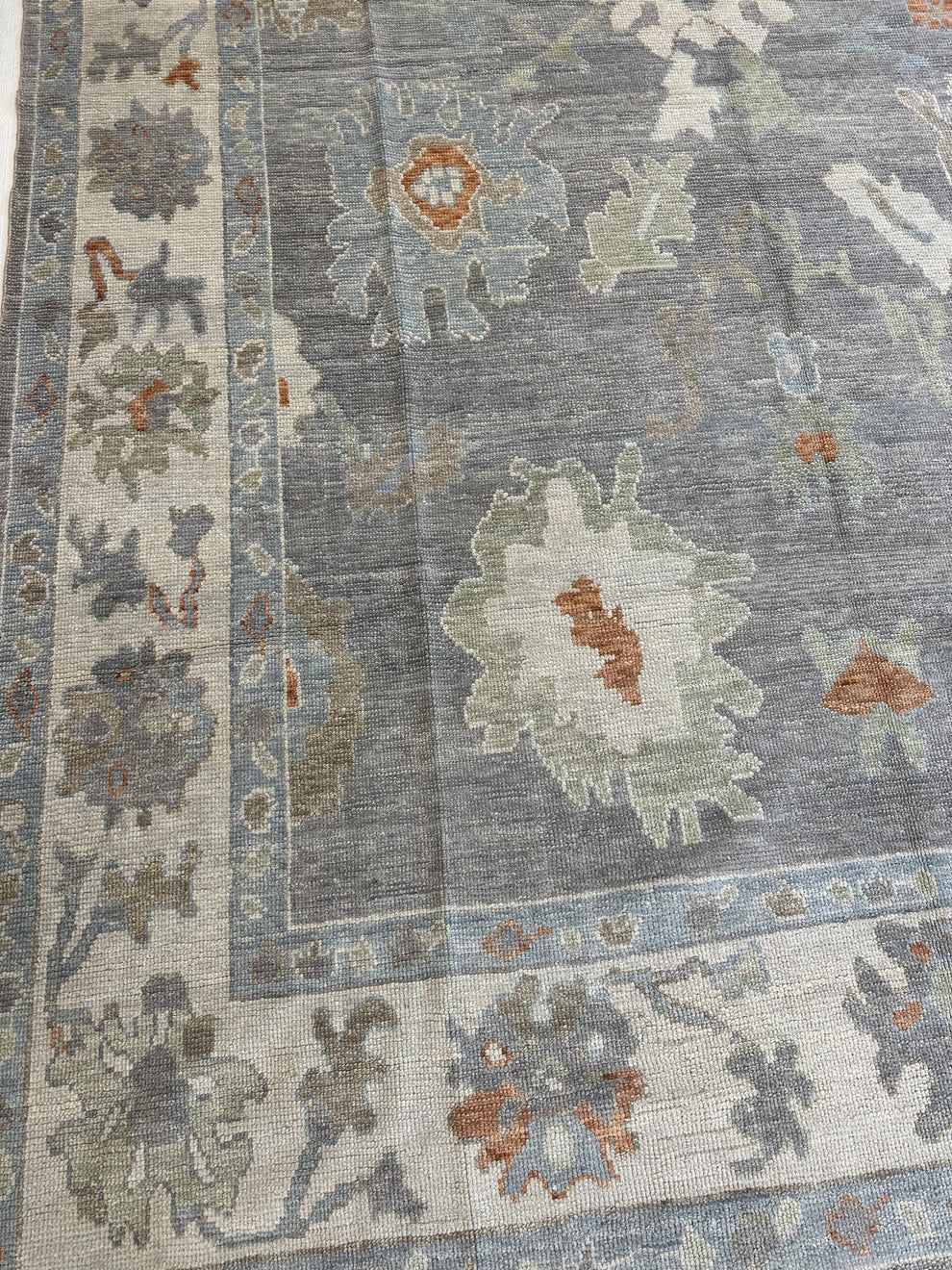 A close-up of a traditional area rug featuring a distressed gray field with floral motifs in muted blue, beige, and rust orange, framed by a detailed floral border.