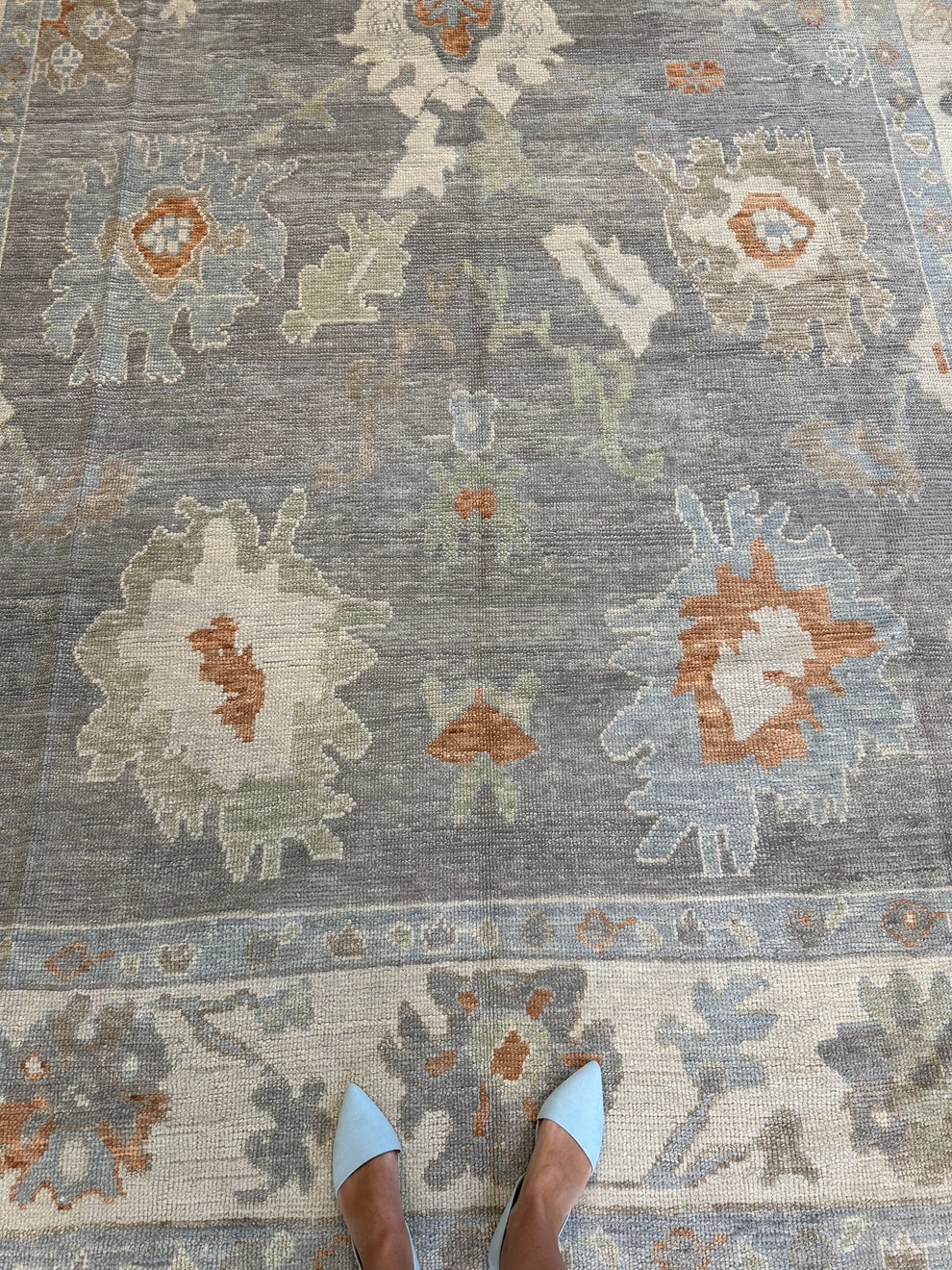 A top-down view of a distressed area rug with a gray base and faded floral motifs in beige, light blue, and rust orange. A person's feet in light blue pointed flats are visible at the bottom edge for scale.