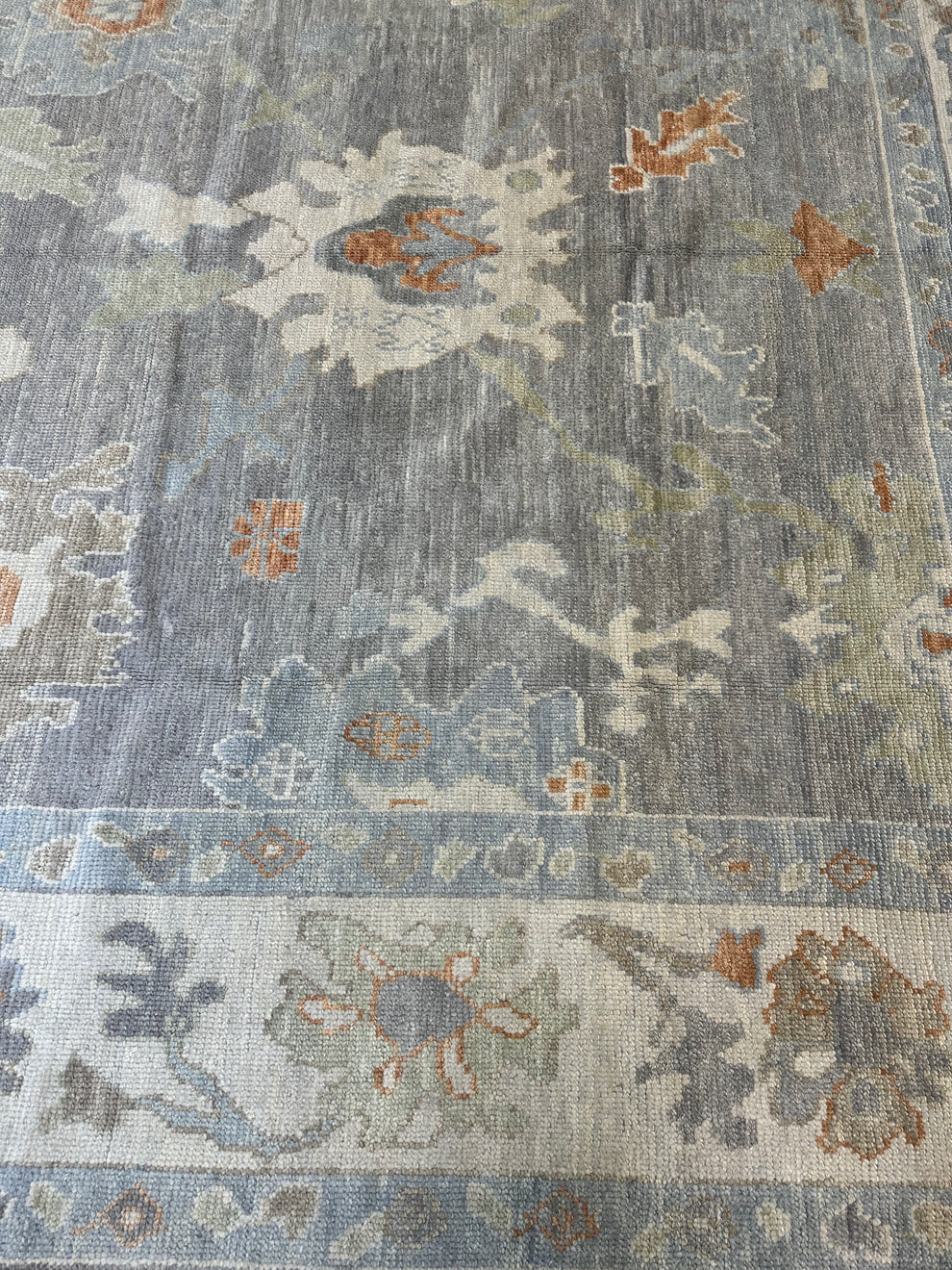 A close-up of a traditional area rug featuring a distressed, vintage-style floral pattern in muted shades of slate blue, beige, and rust orange.