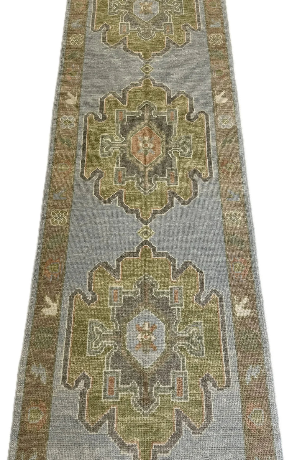 A long rectangular area rug with a traditional geometric medallion pattern in muted olive green, beige, gray, and brown tones, shown flat against a white background.