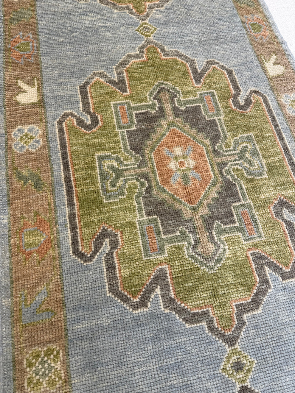 A close-up of a traditional area rug featuring a muted blue-gray field, a large central medallion in olive green, brown, and terracotta, and a patterned border.